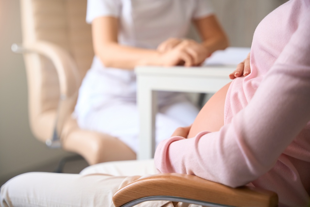 Pregnant woman in a pink sweater sits in a clinic chair while a healthcare professional consults at a nearby desk.