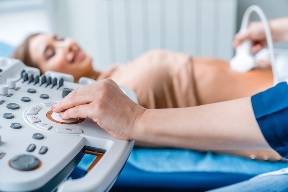 Ultrasound technician using a transducer on a pregnant patient lying on an examination bed with a monitor nearby.
