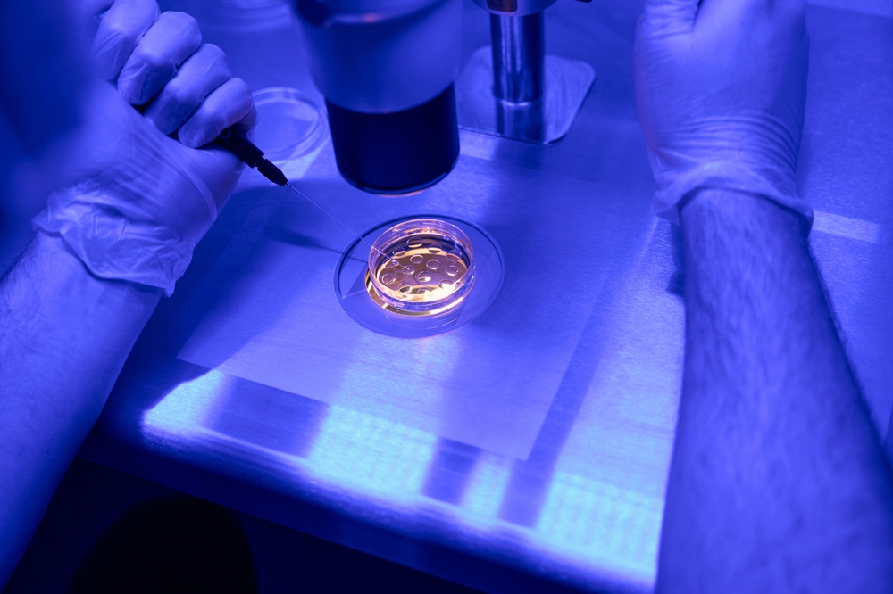 Scientist in gloves uses a microinjection needle toward a petri dish on a blue-lit lab bench under a microscope, performing a delicate procedure