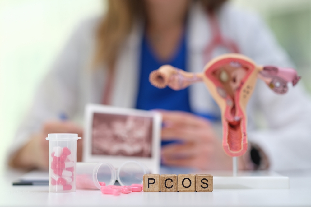 Healthcare professional with a uterus anatomy model and pink pills on a desk; wooden blocks spell PCOS in the foreground
