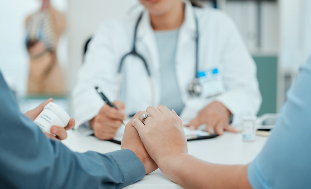 Doctor in a white coat with a stethoscope consults at a clinic as two patients hold hands on the table, signaling support or comfort.