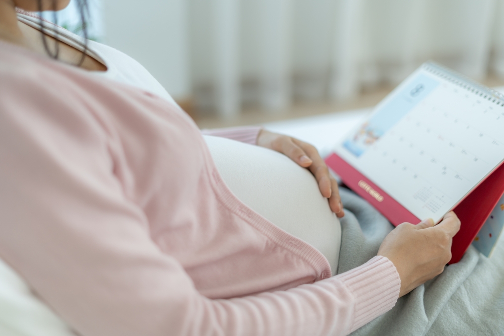 Pregnant person resting with hands on her baby bump while looking at a wall calendar on her lap.