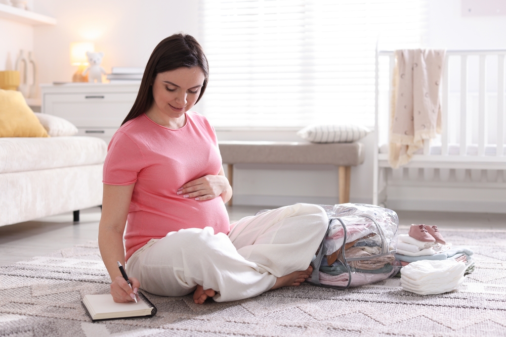 Pregnant woman sitting on the living room floor, writing in a notebook while packing a suitcase with baby clothes nearby.