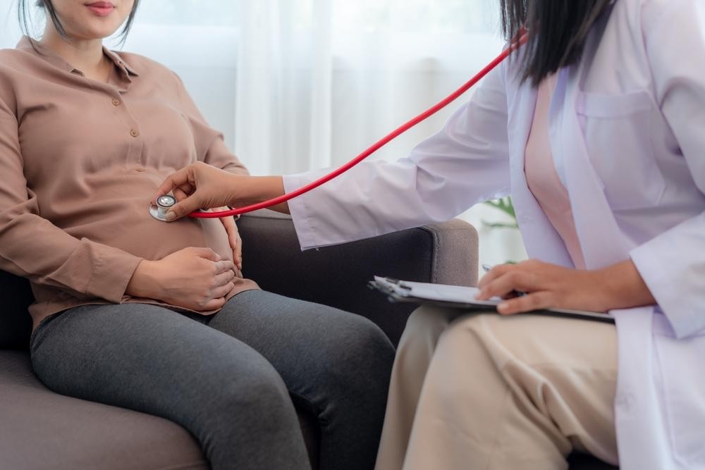 p1 Pregnant woman on a couch while a doctor uses a stethoscope on her belly for a prenatal checkup, doctor taking notes on a clipboard.